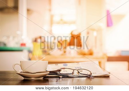 Closeup cup of hot cappuccino coffee with newspaper and spectacles on the wood table with blur coffee shop background-warm tone.