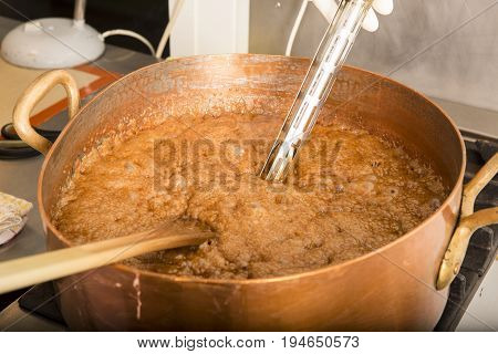 Factory of the production of delicious caramel candies. First step. Boiling syrup of sugar in copper pot with thermometer and big spoon. Brown colour for the syrup.