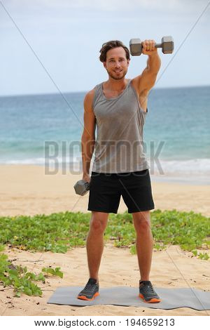 Fitness man lifting dumbbells on beach doing Front Dumbbell Raise i.e. Alternating Front Raise workout for shoulders. Exercising male fitness gym model working out in summer outdoors.