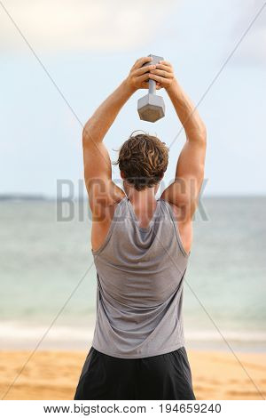 Fitness exercise with dumbbells - Man doing Standing Dumbbell Triceps overhead Extension exercises. Fit male fitness model workout outside on beach gym.