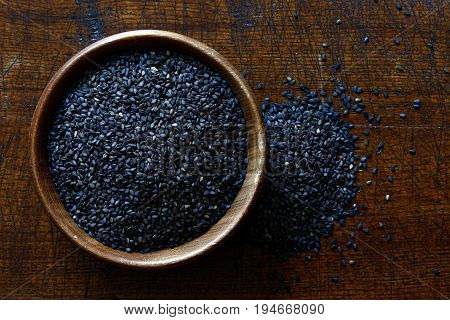 Black Sesame Seeds In Dark Wooden Bowl Isolated On Dark Brown Wood From Above. Spilled Sesame.