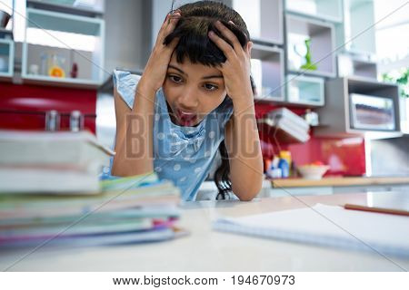 Shocked girl with head in hands by books at kitchen counter