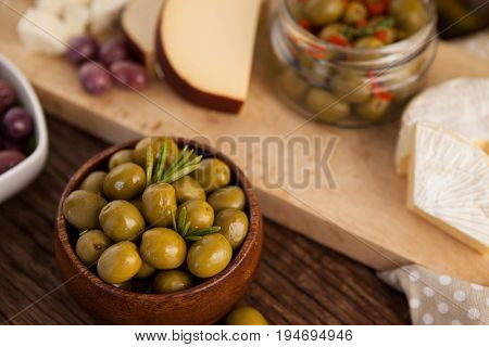 Green olives in container by cheese and vegetable on cutting b oard