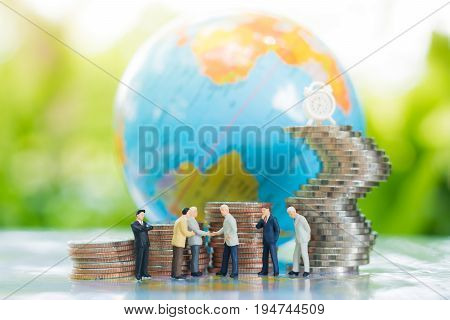 Miniature businessman handshaking with globe and growing stacks of coins background using as commitment agreement investment and partnership business concept.