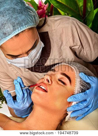Filler injection for female lips augmentation. Face plastic aesthetic facial surgery in beauty clinic. Doctor in medical gloves with syringe injects lips augmentation. Green plants in background.