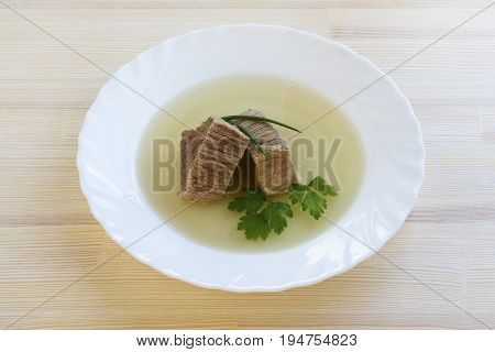 Homemade beef broth or bouillon with pieces of beef in a white plate on a wooden table view from above.