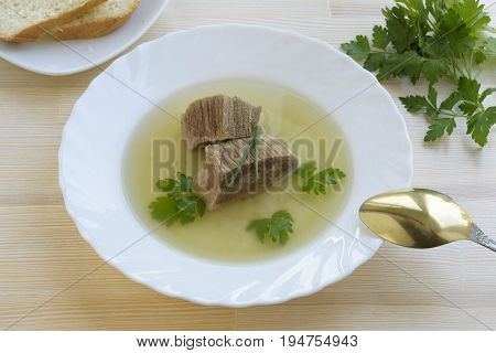 Homemade beef broth or bouillon with pieces of beef in a white plate with golden spoon on a wooden table view from above.