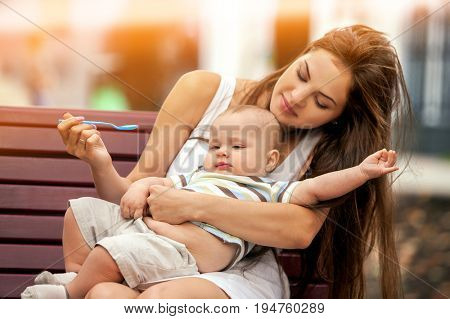 Baby feeding spoon by mother in park outdoor. Weaning in first weeks. Mom and eating child summer on bench. Good appetite by baby food with foster mother . Color tone on shiny sunlight background.