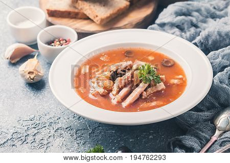 Soup with meat, olives, herbs, lemon in white bowl, black bread and spices on gray background, homemade food. Traditional Russian soup - solyanka