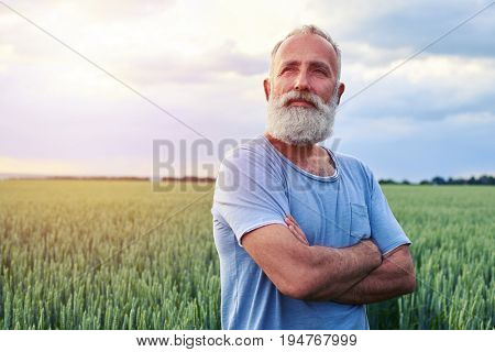 Close-up shot of beard mature man with folded arms, standing in the green field and looking somewhere 