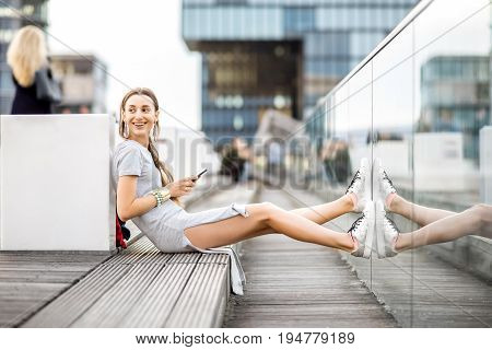 Lifestyle portrait of a business woman dressed casually sitting with phone outdoors at the modern district in Dusseldorf city, Germany