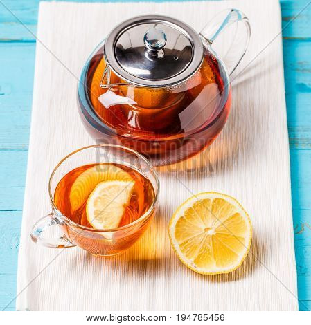 Glass cup of tea with lemon and glass teapot on blue wooden table.