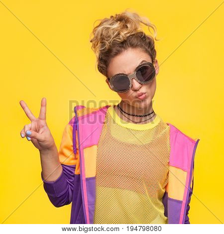 Blonde hipster girl with hair bun wearing colorful jacket, yellow sheer shirt grimacing making v-sign.