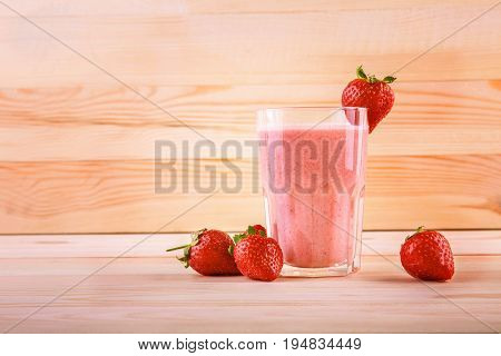A glass of fresh, tasty, sweet and beautiful smoothies on a light brown wooden table. One strawberry is on top on a glass cup. A few strawberries are near a full jar of strawberry drink.