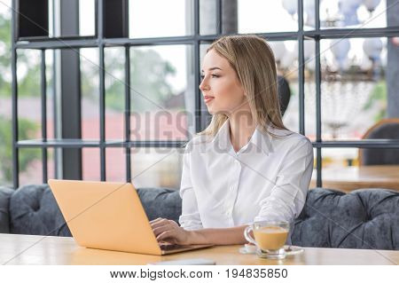 Gorgeous young happy female blond with straight hair sits on the sofa and looks away. Portrait of beautiful and successful business woman in white blouse and with laptop on the light brown table.