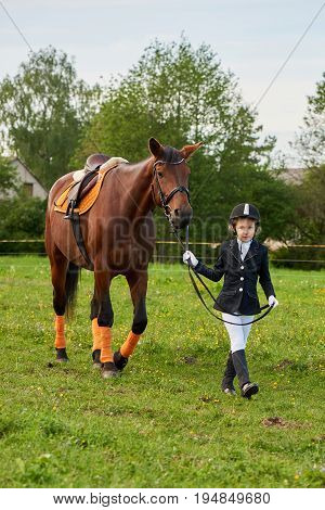 Pretty little girl jockey lead horse by its reins across country in professional outfit