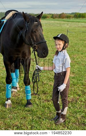 Pretty little girl jockey communicating with her black horse in professional outfit