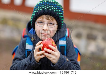Little kid boy with eye glasses on the way to the school and eating fresh apple. Child on a street. Preschooler wearing warm clothes, glasses and backpack. Happy student or people. Education concept