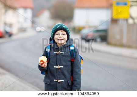Little kid boy with eye glasses on the way to the school and eating fresh apple. Child on a street. Preschooler wearing warm clothes, glasses and backpack. Happy student or people. Education concept