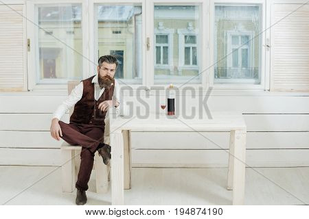 Hipster Sitting On Chair At Table With Bottle And Glass