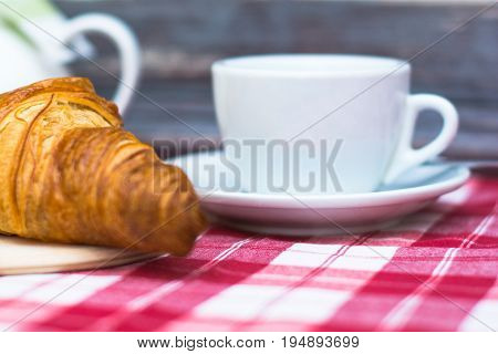 Fresh croissant and a white cup of coffee on a red white checkered tablecloth near a wooden wall. French Breakfast. Coffee break.