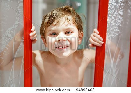 Happy little kid boy in the shower cabin wet and smiling. Adorable child with blond hairs taking bath and having fun.
