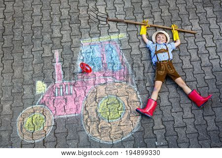 Happy little kid boy in straw hat and rain boots having fun with tractor picture drawing with colorful chalks. Children, lifestyle, fun concept. child dreaming of future and profession.