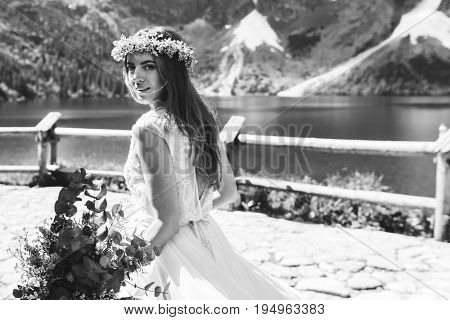 the bride in a beautiful dress and a wreath of white flowers holding a bouquet of flowers and eucalyptus overlooking the mountains and lake