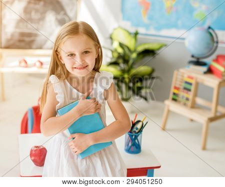 Little smiling blond girl standing near blackbord with red backpack and holding a notebook with pencils in the sunshine school classroom