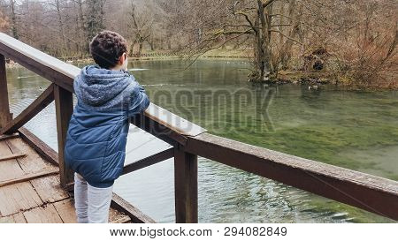 Young Boy Standing On The Wooden Bridge And Enjoying The Picturesque View Of The River.
