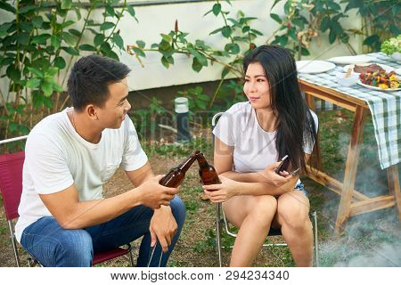 Smiling Young Asian Cuople Clinking Beer Bottles When Grilling Meat In Backyard