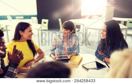 education, high school, people and technology concept - group of international students sitting at table with tablet pc computers, books and notebooks talking at university