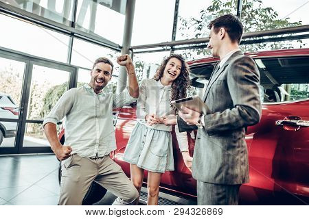 Sales Situation In A Car Dealership, The Dealer Is Handing Auto Keys To A Young Couple, They Are Exc