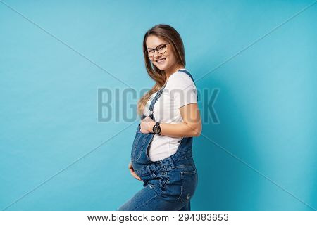 Pregnant Woman Caressing Her Belly Over Blue Background. A Cute Young Woman Wearing Glasses, And Den