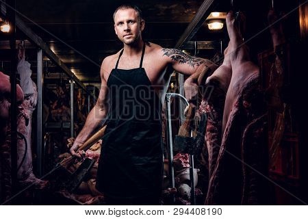 Shirtless Butcher With A Tattoo On His Hand Wearing Apron Posing With Two Axes In A Refrigerated War