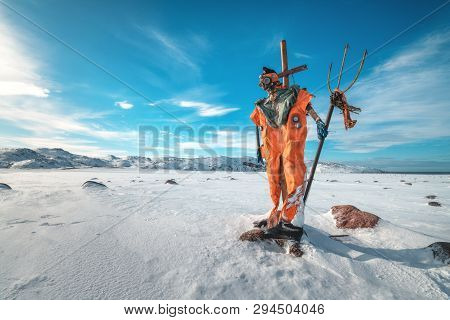 Scarecrow In Orange Clothes And Gas Mask With Trident Is Against Blue Sky With Clouds And Snowy Mead