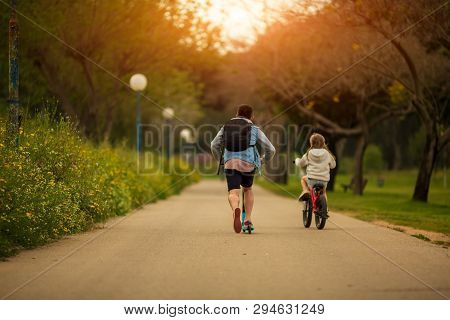 The Father With The Daughter Walk In The Park On A Scooter And Bike In Sunny Summer Day
