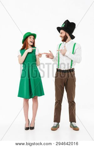Full length of a happy young couple wearing costumes, celebrating St.Patrick 's Day isolated over white background, pointing fingers at each other
