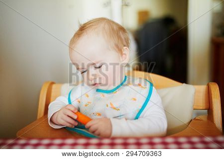Cute Baby Girl Eating Carrot In The Kitchen. Little Kid Tasting Solids At Home. Baby Led Weaning, He
