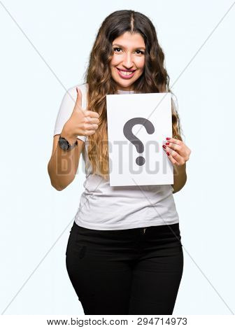 Young adult woman holding paper with question mark happy with big smile doing ok sign, thumb up with fingers, excellent sign