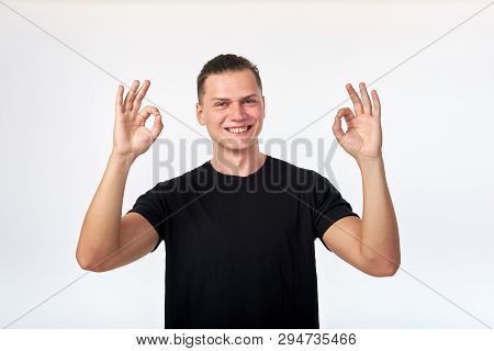 Attractive Cherrful Young Man Dressed In Black Shirt Showing Ok Gesture, Smiling And Looking At Came