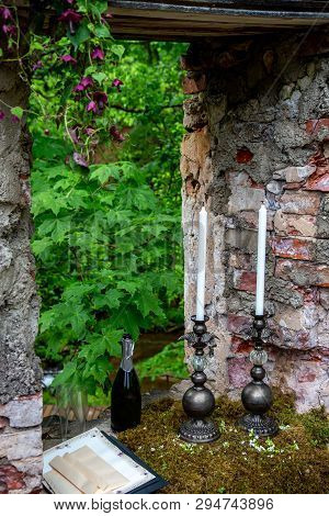 Candles In A Candlestick, Bottle Of Champagne And Two Glasses In Moss At Stone Ruins. Wedding Celebr