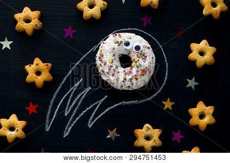 Donut Meteorite With Googly Eyes Among The Stars Of Cookies On A Black Chalkboard Background, Funny 