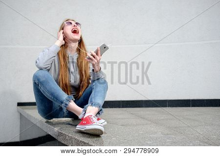 Redhead Girl Dressed In Hoody, Jeans And Red Sneakers, With Red Lipstick Painted Lips Listening To M