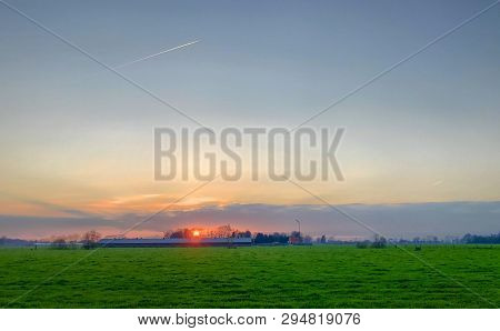 Dramatic And Colorful Sunrise Or Sunset Sky Over A Grassy Green Farmfield Lined By Trees