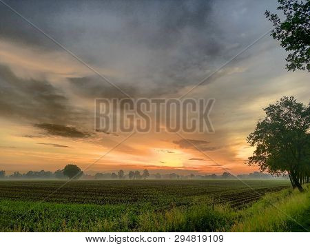 Dramatic And Colorful Sunrise Or Sunset Sky Over A Grassy Green Farmfield Lined By Trees
