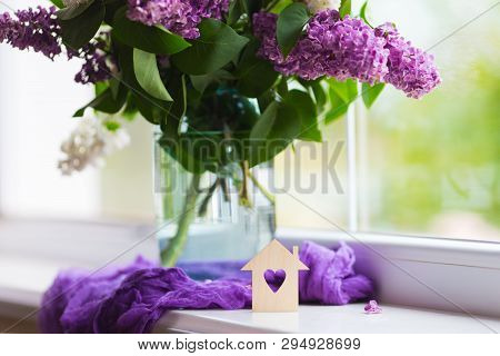 Wooden House With Hole In Form Of Heart And Tender Bouquet Of Beautiful Lilac In Glass Vase Near Win