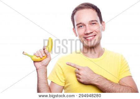 Smiling Handsome Guy Holding Yellow Banana And Pointing To It Over White Background. Studio Shot.