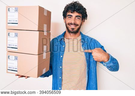 Handsome young man with curly hair and bear holding delivery package pointing finger to one self smiling happy and proud 