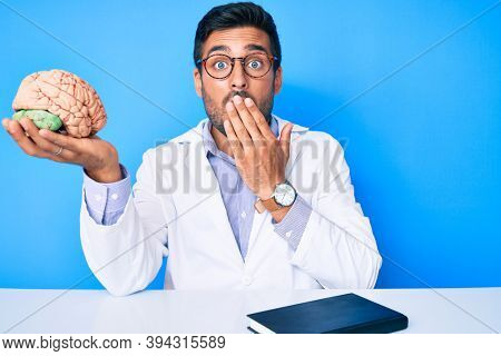 Young hispanic man sitting at the table wearing doctor coat holding brain covering mouth with hand, shocked and afraid for mistake. surprised expression 